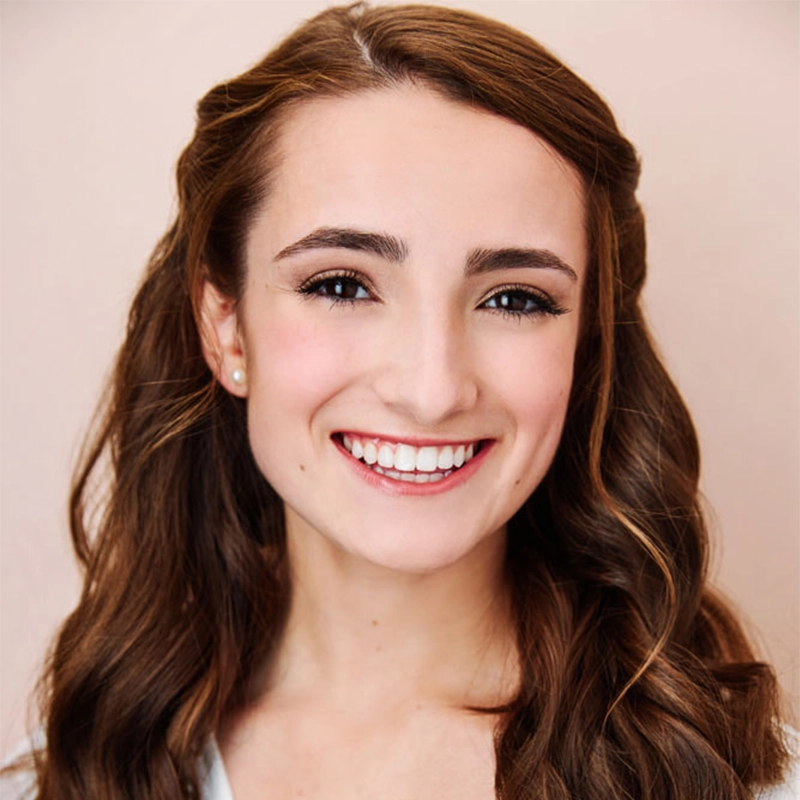 Alice K. Tinari, a young woman with long, wavy brown hair, smiles warmly at the camera. She has fair skin, brown eyes, and is wearing natural makeup with light pink lipstick. She is dressed in a white top against a soft beige background.