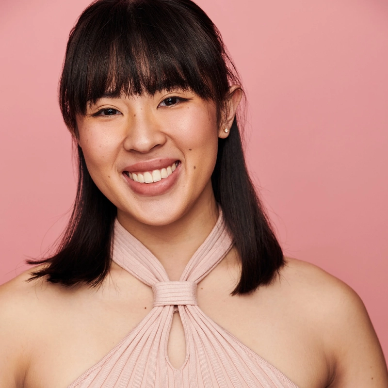Regine Sophia, a young woman with straight, shoulder-length dark hair and bangs, smiles warmly. She wears a sleeveless, light pink halter top with a keyhole neckline against a solid pink background for a cheerful, cohesive palette.