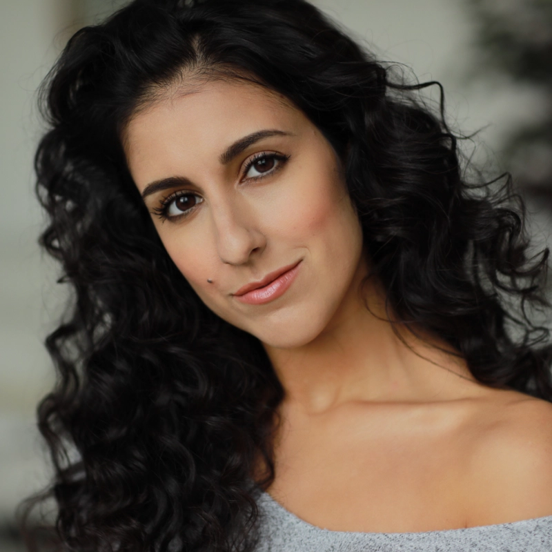Gabriella Sorrentino, with long, dark, curly hair and light skin, smiles softly at the camera. She wears natural makeup, light pink lipstick, and a grey off-the-shoulder top against a softly blurred background.