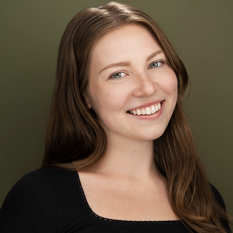 A young woman with long brown hair and fair skin smiles warmly at the camera. She is wearing a black top with a square neckline. The background is a soft olive green, creating a calm and pleasant atmosphere.