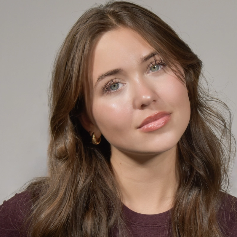 Lilly Mae Stover, a young woman with long, wavy brown hair and light green eyes, looks slightly to the side with a soft, relaxed expression. She wears gold hoop earrings, a dark top, and natural makeup against a plain light gray background.