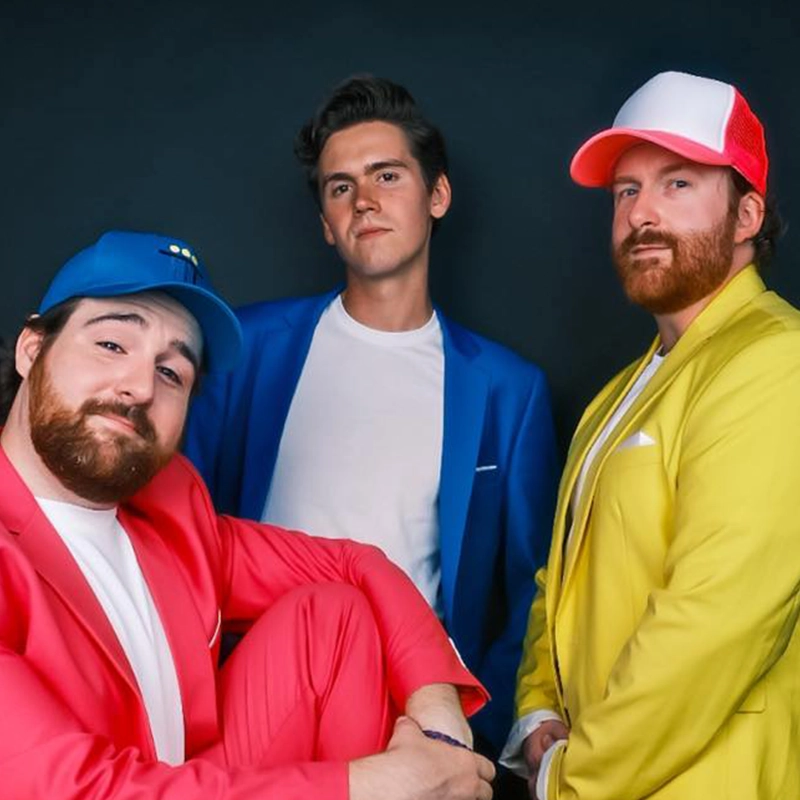 Three men pose together against a dark background. One wears a pink suit and blue cap, another wears a blue suit with a white shirt, and the third wears a yellow suit and red-and-white cap. All have facial hair and neutral to confident expressions.