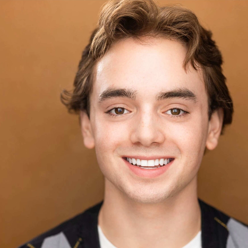 Sophia Leverett, a young person with light skin, short brown hair, and brown eyes, smiles directly at the camera. The background is a warm, solid tan color. She wears a dark top over a white shirt under bright, even lighting.