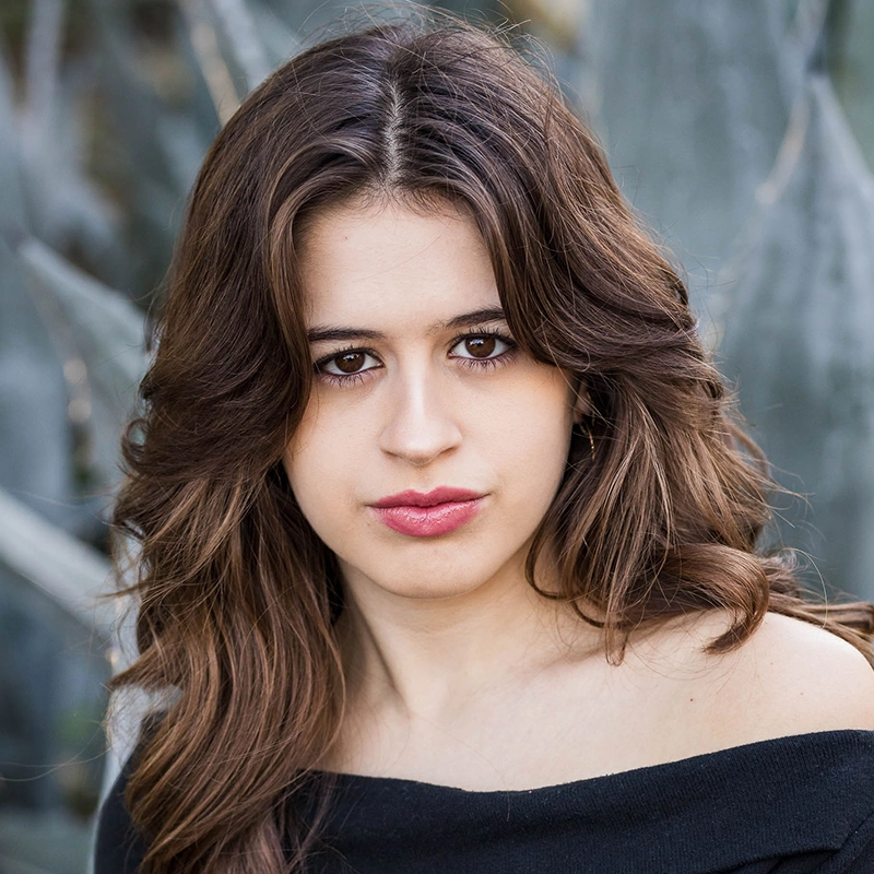 A young woman with wavy brown hair and brown eyes looks directly at the camera. She wears an off-shoulder black top. The background is blurred with hints of green and gray, possibly from plants or trees—an image reminiscent of the expressive portraiture style favored by Timothy Thomas. Her expression is neutral, lips slightly pursed.