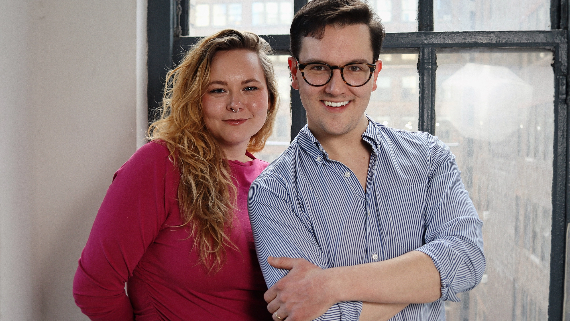 The Voice Collective site image 26 Two people stand smiling in front of a large window. The person on the left has long, wavy blonde hair and wears a pink top. The person on the right, part of The Voice Collective, has short brown hair, glasses, and a blue striped shirt with rolled sleeves.