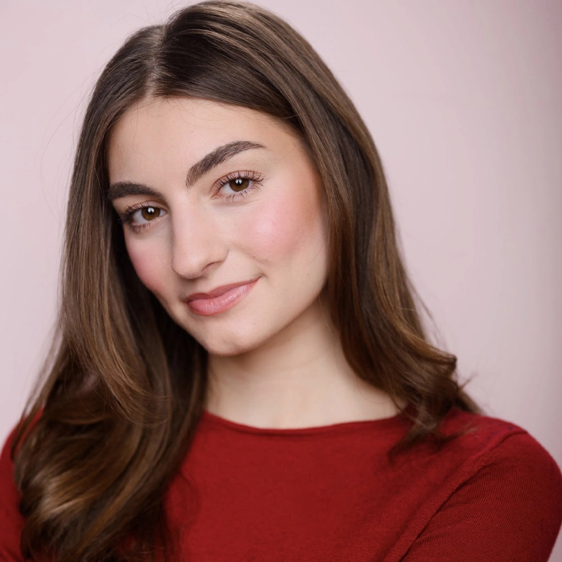 Sophia Theokas, a young woman with long brown hair and fair skin, smiles softly at the camera. She wears a red top and has natural makeup with pink blush against a soft, pale pink background for a warm, inviting feel.