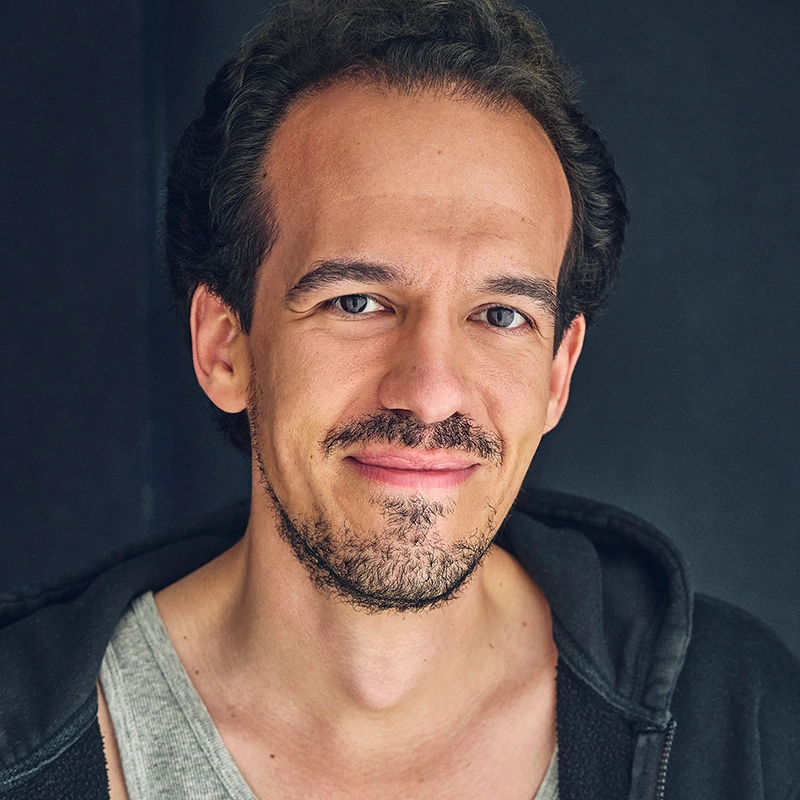 A man with short dark hair and facial hair smiles gently at the camera. He is wearing a gray tank top and a black hoodie. The background is plain and dark, making Thomas Vorsteg’s face and casual attire stand out. The lighting is soft and even.