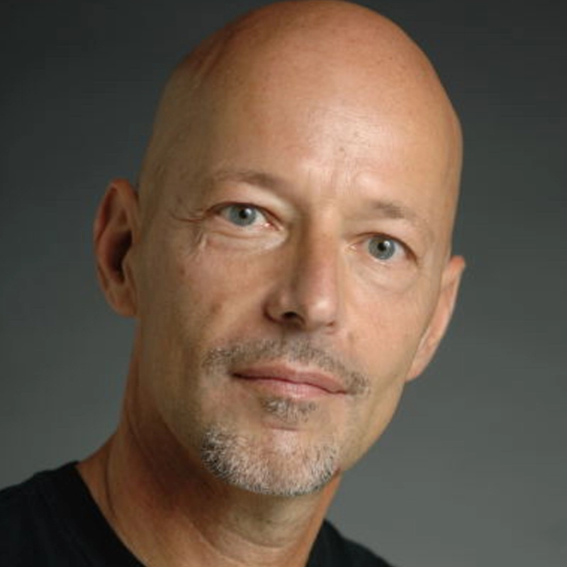A middle-aged bald man with light skin, blue eyes, and a trimmed salt-and-pepper goatee looks at the camera with a neutral expression. He wears a black shirt and is set against a plain, dark background.