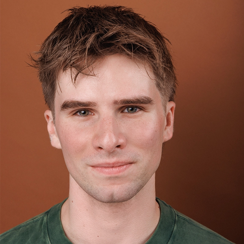A young man with short brown hair wears a green shirt and smiles slightly. He has fair skin, blue eyes, and neatly groomed eyebrows. The background is a plain, warm brown color. The lighting is soft, highlighting his facial features. This could easily be a portrait of Timothy Thomas.
