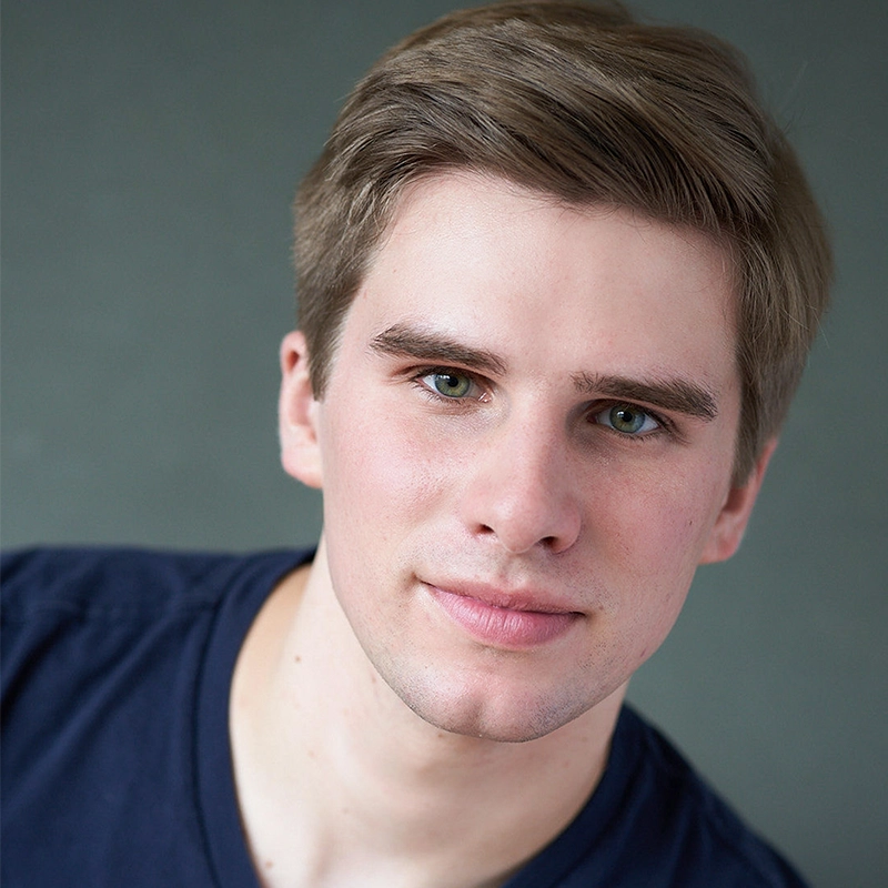 A young man with fair skin and short, light brown hair looks directly at the camera. He has green eyes and a slight smile. He is wearing a dark blue T-shirt and is posed against a plain, muted gray background.