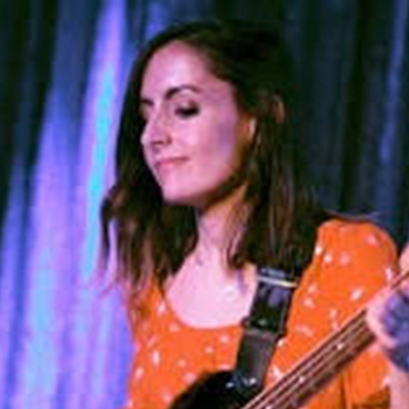 A woman with shoulder-length brown hair plays an electric guitar. She is wearing an orange top with white patterns and is standing in front of a purple curtain. Her eyes are closed, and she appears to be focused on the music.