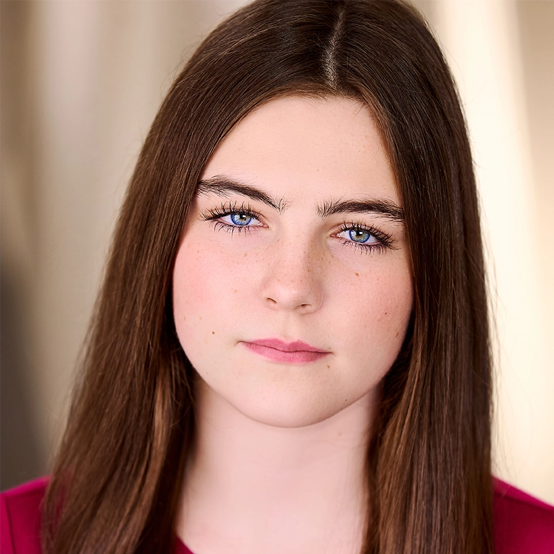A young woman with long brown hair and bright blue eyes looks directly at the camera with a neutral expression. Jackie Winslow has lightly freckled, fair skin and wears a dark pink top, set against a softly blurred beige background.