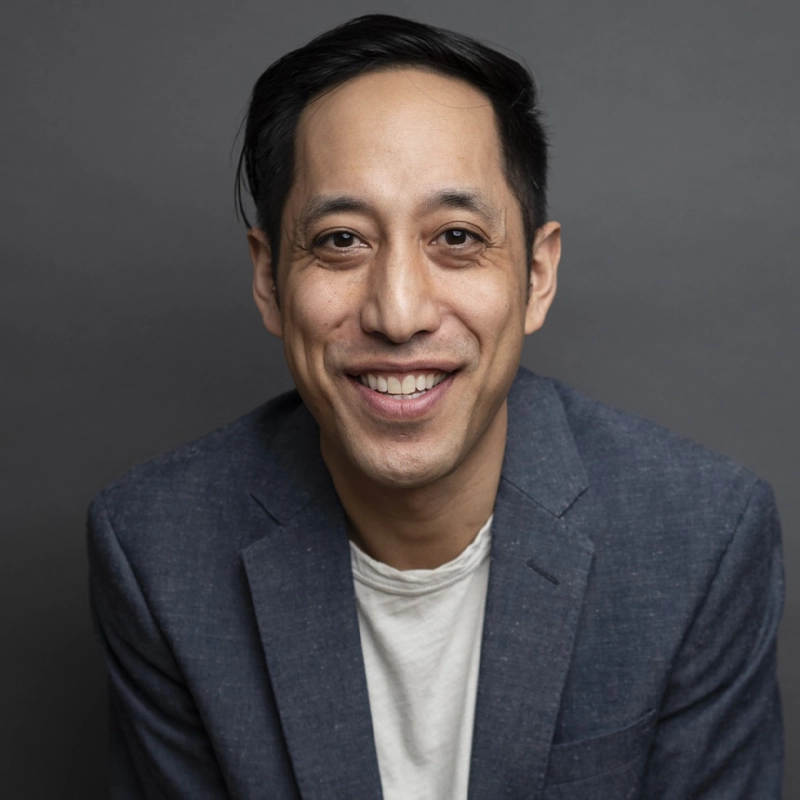 A man with straight dark hair and a side part, wearing a gray blazer over a white shirt, smiles at the camera in front of a plain gray background.