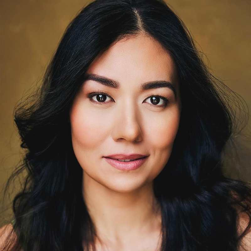 Isabelle Van Vleet, with long wavy black hair and warm-toned skin, faces the camera smiling softly. She has defined eyebrows, brown eyes, and natural makeup with pink lipstick against a plain, warm brown background.