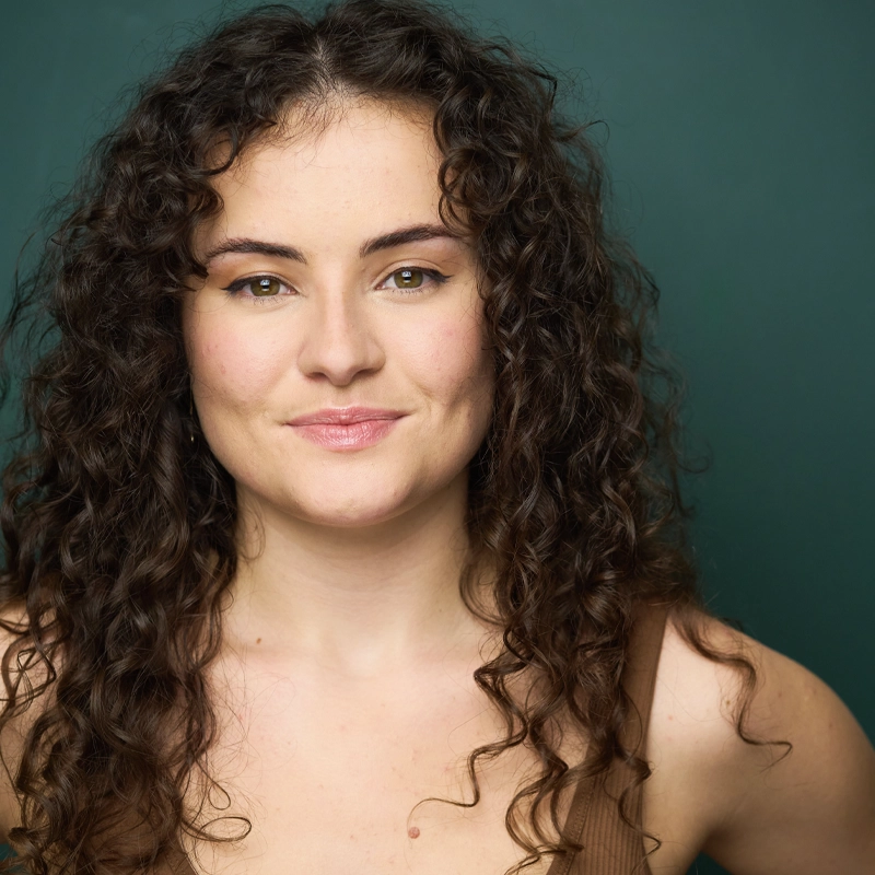 Delaney White, with long, dark, curly hair and light skin, smiles softly at the camera. She wears natural makeup and a brown sleeveless top against a solid dark green background. Her expression is warm and confident.