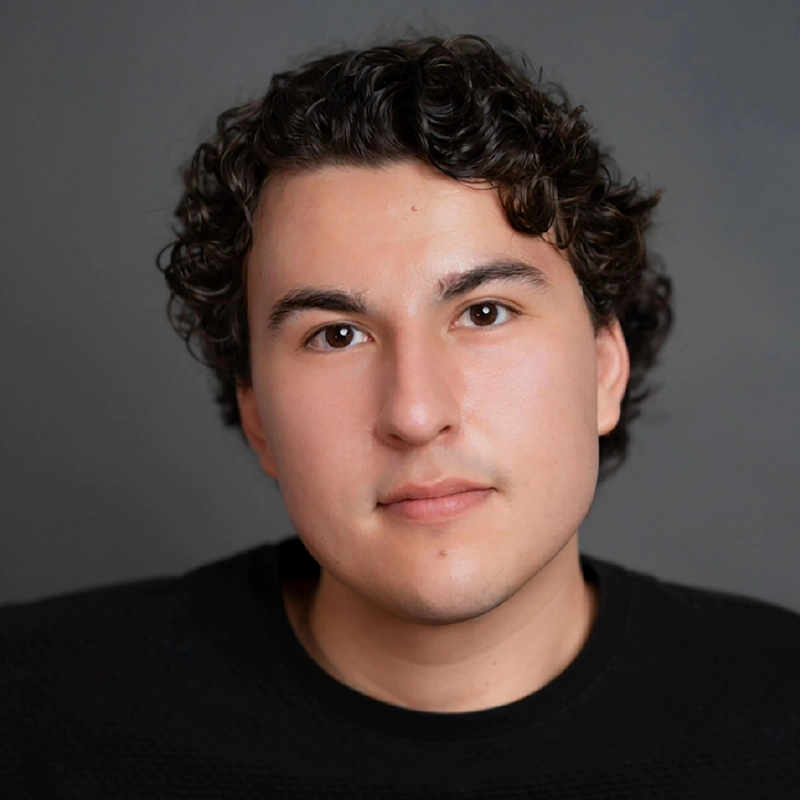 A young person with short, dark, curly hair and fair skin looks directly at the camera with a neutral expression. They are wearing a black top and are set against a plain, dark gray background. The lighting is soft and even.