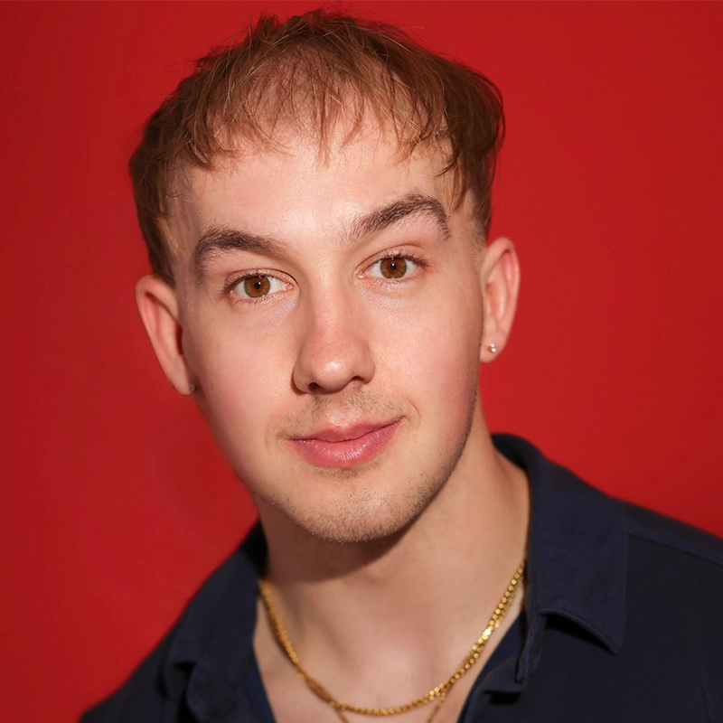 A young person, identified as Timothy Thomas, with short, light brown hair and brown eyes is facing the camera, smiling slightly. They wear a navy blue shirt, a gold chain, and an earring. The background is solid bright red.