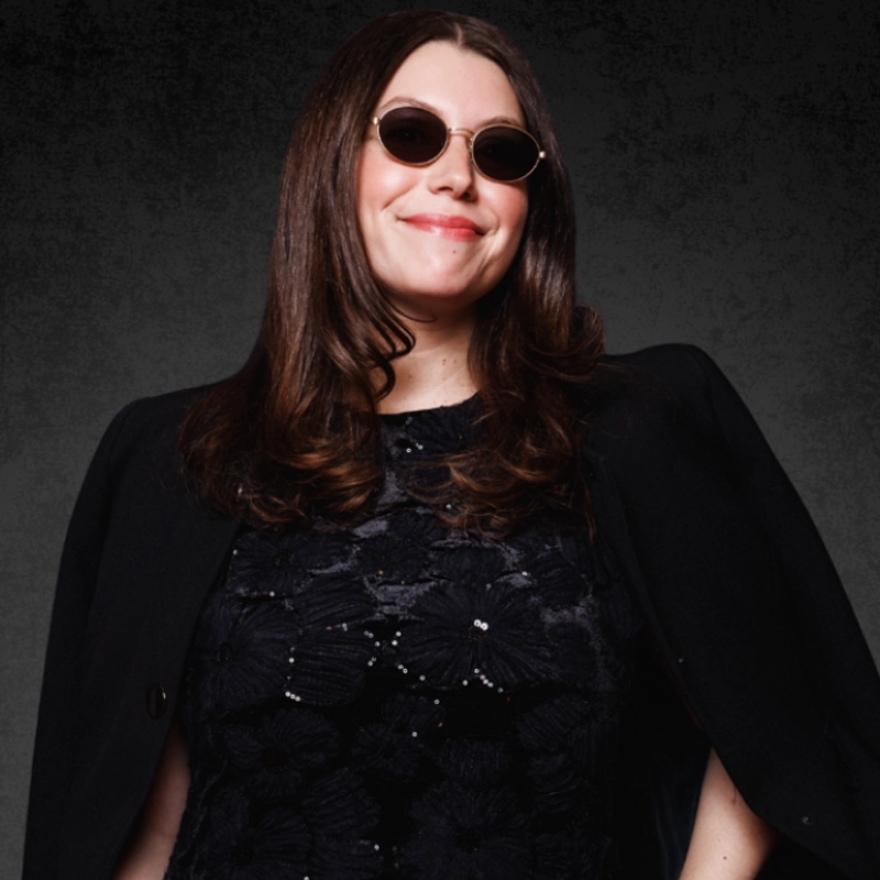 Lindsay Wormser, with long brown hair, wears round sunglasses and a black textured dress with a matching black coat draped over her shoulders. She smiles brightly while standing against a dark, subtly textured background.
