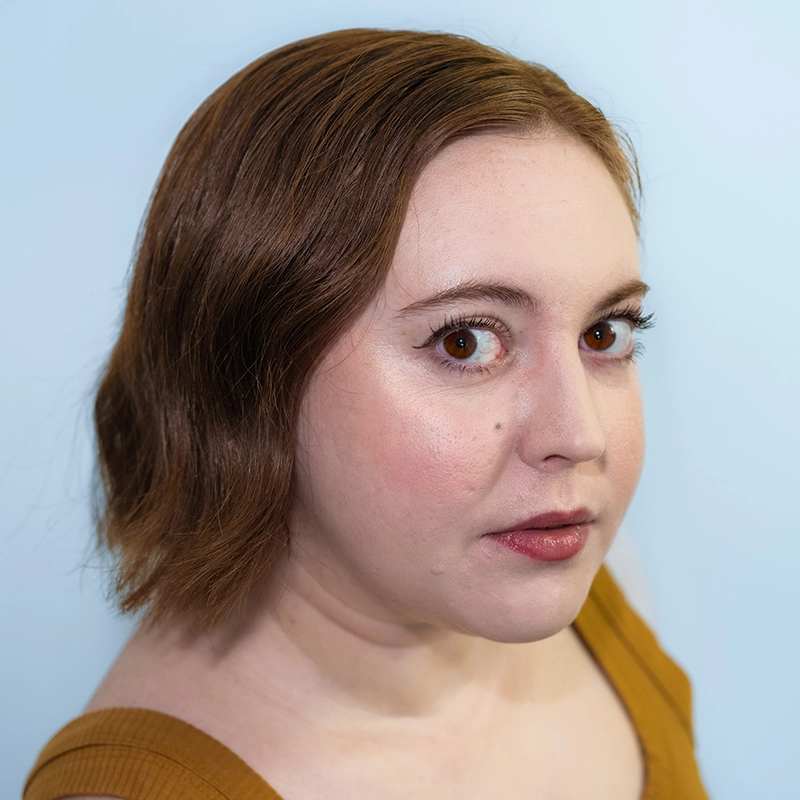 A person with fair skin, brown wavy shoulder-length hair, and bold eyeliner looks at the camera against a plain light blue background. They wear a mustard-colored sleeveless top. Their expression is neutral, with lips slightly parted and eyes wide—a look reminiscent of Timothy Thomas’s understated yet expressive style.