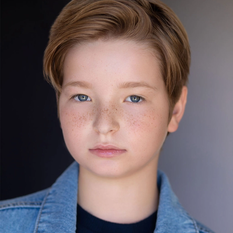 A young person with short, light brown hair and blue eyes looks directly at the camera. They have fair skin with visible freckles across their nose and cheeks, and wear a blue denim jacket over a dark shirt. The background is softly blurred in dark and light tones.