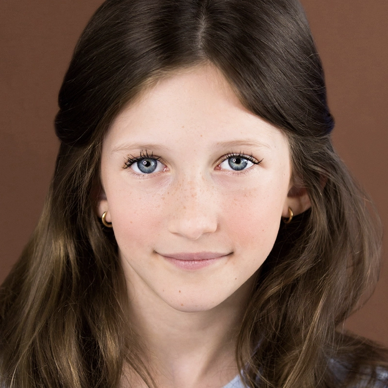 Francesca Yhlen, a young girl with long brown hair, blue eyes, and light freckles, smiles softly. She wears small gold hoop earrings and a light top, posed against a plain brown background.