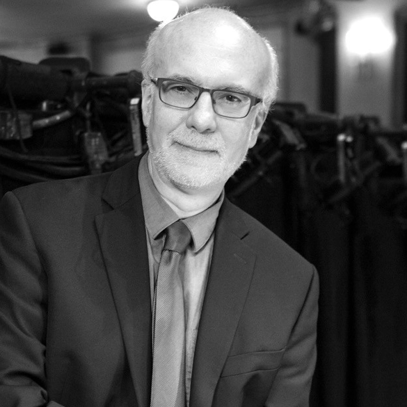 A middle-aged man, Larry Yurman, with glasses, short white hair, and a beard is pictured in black and white. He smiles gently in a suit jacket, dress shirt, and tie while standing indoors before dark drapes and lighting equipment.