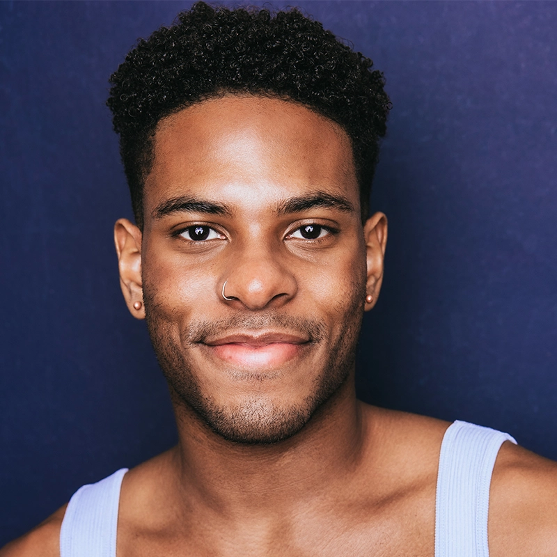A young man with short curly hair smiles softly at the camera. He wears a nose ring, small hoop earrings, and a light purple tank top by Maggie Weller. The solid dark blue background highlights his relaxed, friendly expression.
