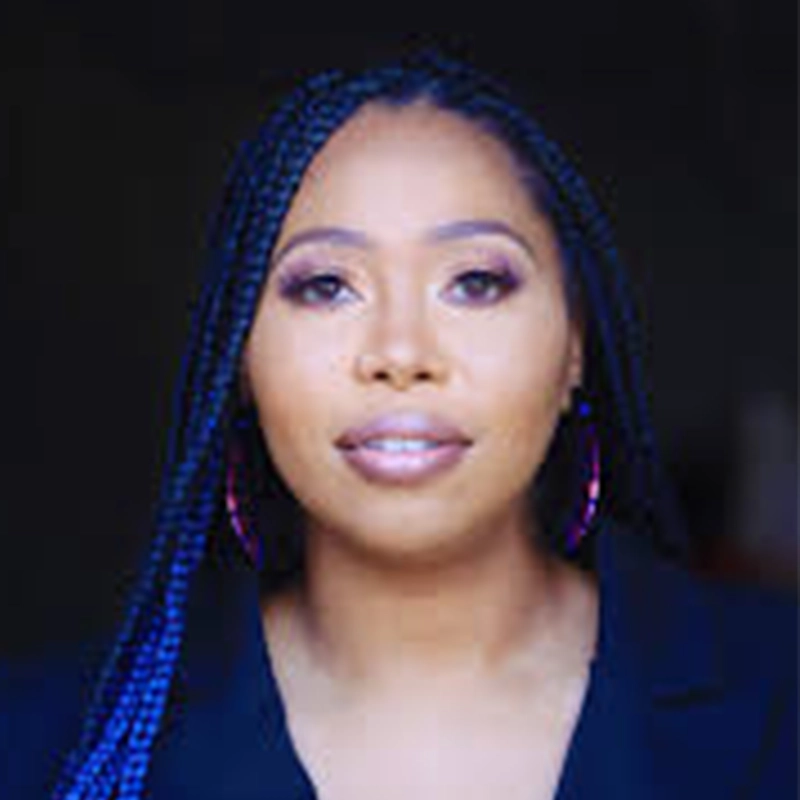 A woman with long, braided hair wears hoop earrings and a black top. She has natural makeup with defined eyebrows and nude lipstick, looking directly at the camera against a dark, blurred background—resembling Amanda Quaid.