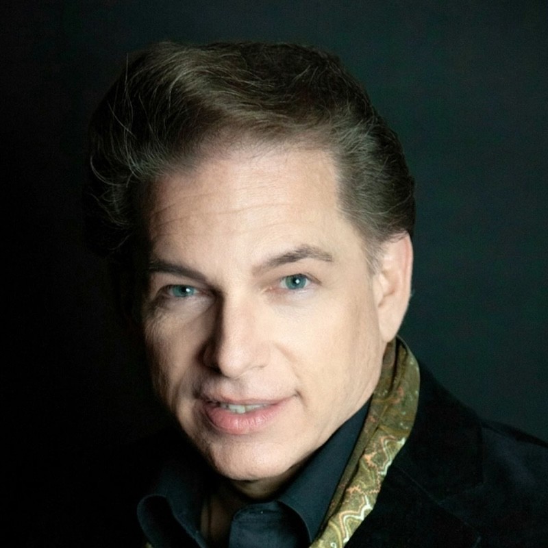 A man with light skin, blue eyes, and styled brown hair smiles at the camera. Dorian Woodruff wears a black shirt, a black jacket, and a paisley-patterned scarf, with a dark background behind him.