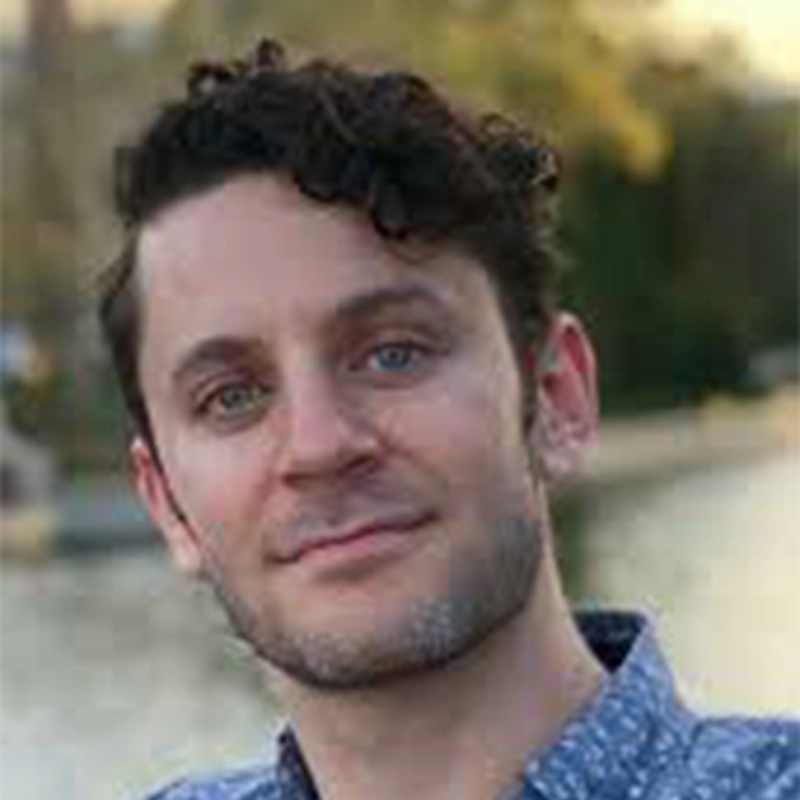 Garrett Bell, a man with light skin, short curly brown hair, and blue eyes, smiles gently at the camera. He has light facial stubble and wears a blue patterned shirt. The soft-focus background shows water and green trees in daylight.