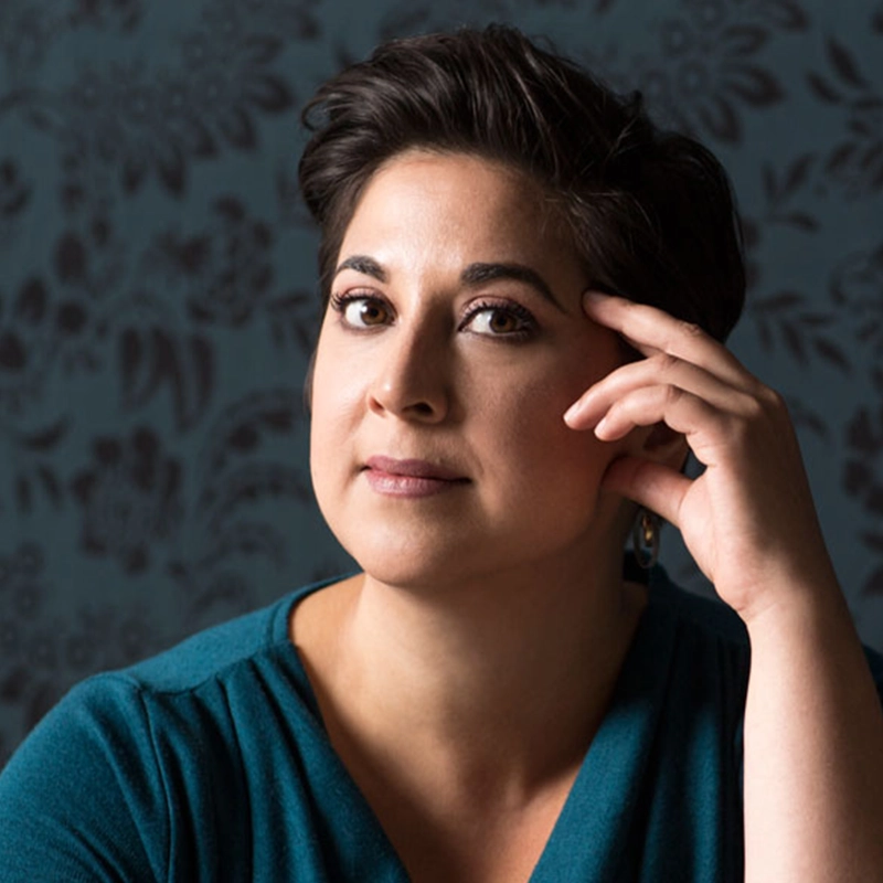 Amanda Quaid, with short dark hair and brown eyes, gazes thoughtfully at the camera, resting her head on her hand. She wears a teal top before a dark floral-patterned background. Soft lighting highlights her face.