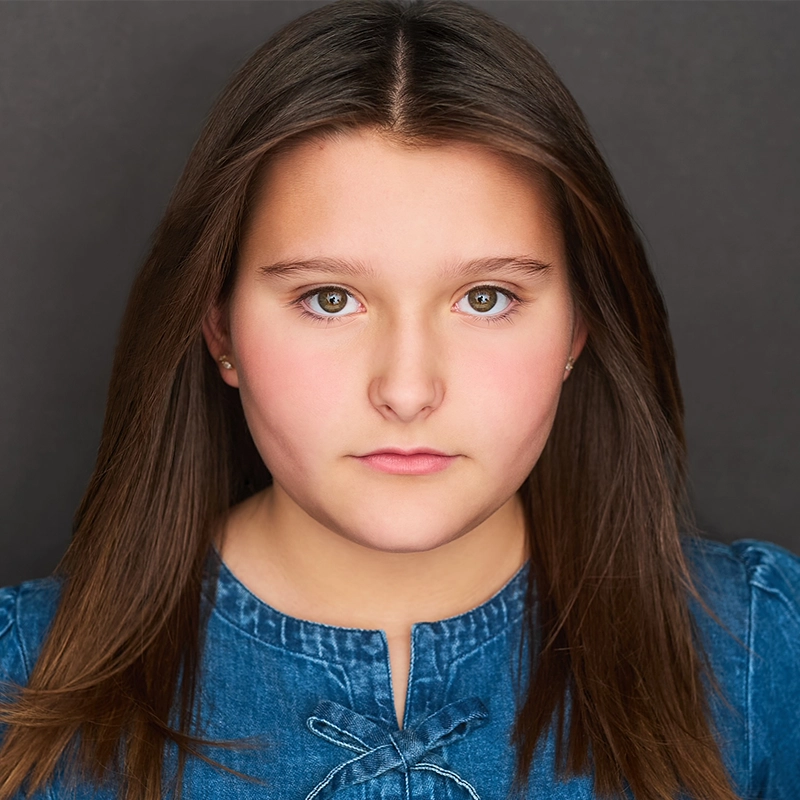 A young girl with straight brown hair and fair skin stares directly at the camera with a neutral expression. She wears a blue denim top with decorative stitching. Jackie Winslow is set against a plain, dark background, drawing focus to her face.