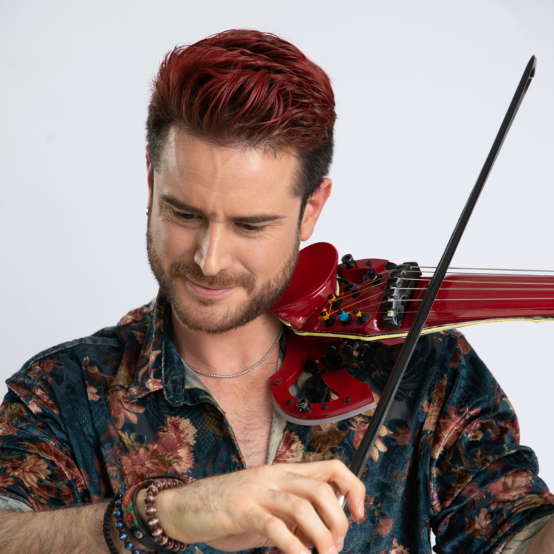 A man with red hair and a trimmed beard plays a red electric violin. He wears a velvet floral shirt and beaded bracelets. His expression is focused, and the background is a plain, light color.