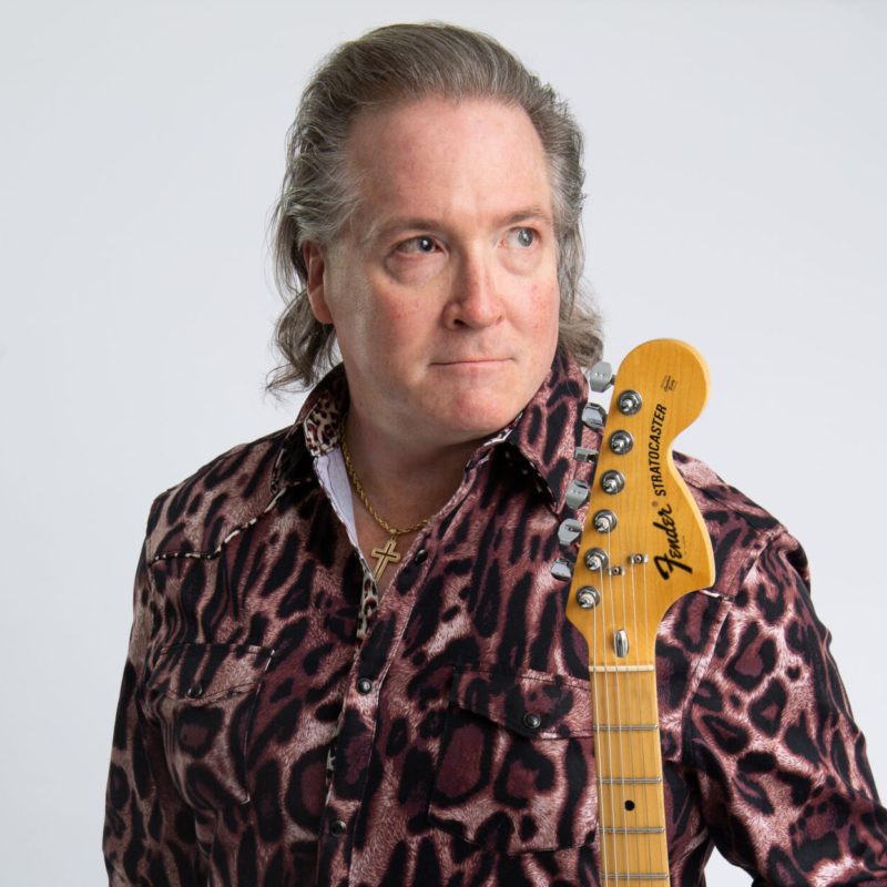 A man with gray-blonde hair and fair skin wears a pink and black animal-print shirt and a gold cross necklace. He holds a Fender Stratocaster guitar over his right shoulder and looks to his left with a neutral expression against a plain light background.