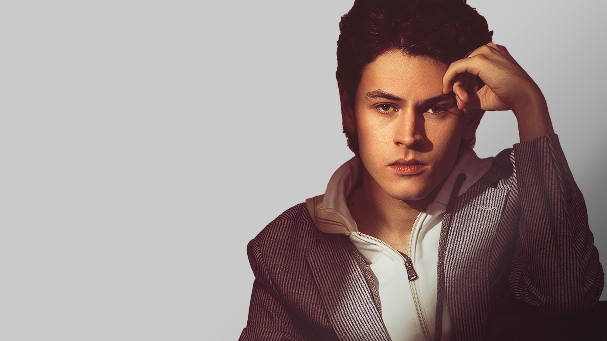 A young man with wavy dark hair sits against a light gray background. He wears a zippered white shirt under a striped jacket. Casey Likes rests his right hand on his forehead, gazing back to the past with a thoughtful, serious expression.