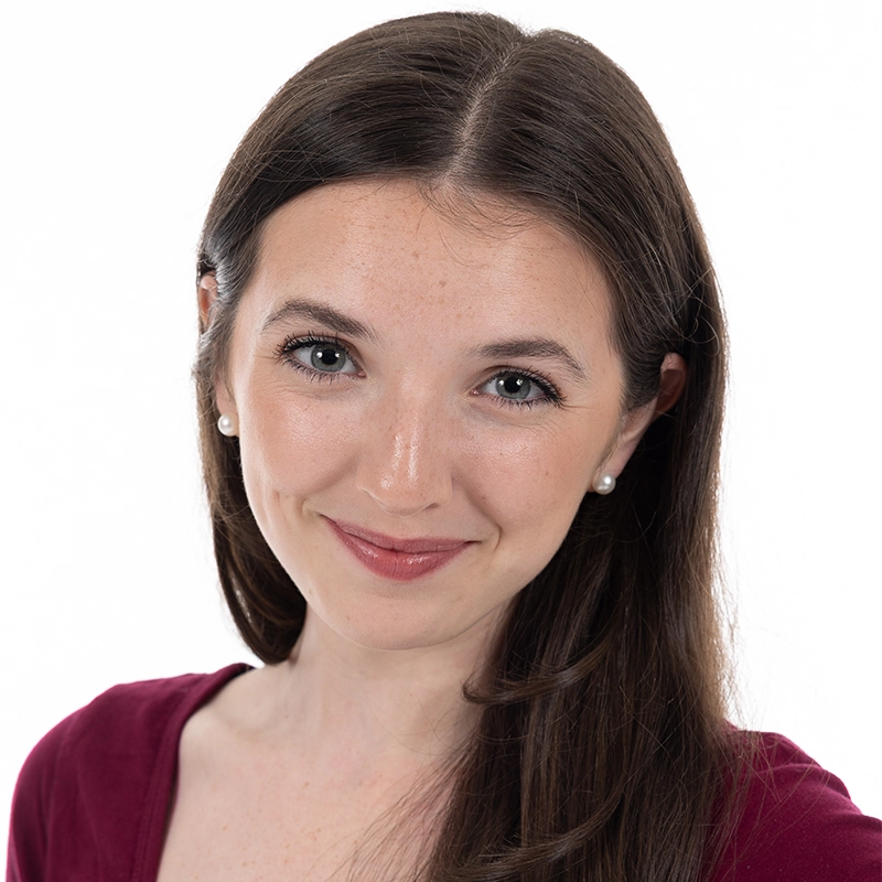 Gabriella Santalla, a young woman with long brown hair and fair skin, smiles gently. She wears pearl stud earrings and a deep red top. Her makeup is natural, with subtle pink lipstick against a bright white background.