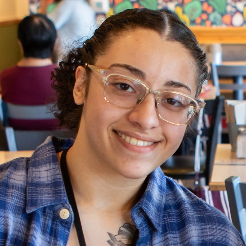Abigail Rodriguez, a young person with glasses, short curly hair, and a plaid shirt, smiles at the camera in a colorful, busy café. People and tables fill the background, and a tattoo is visible on her chest.