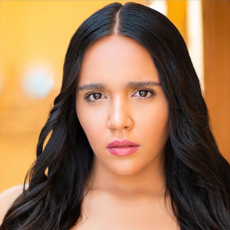 A woman with long, wavy black hair poses against a warm, blurred yellow background. She has light skin and expressive brown eyes. Jett Roberts wears natural makeup with pink lipstick and gazes seriously and directly into the camera.