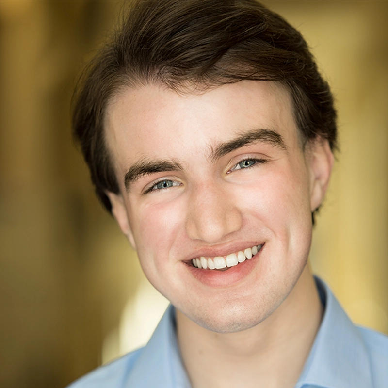 A young man with fair skin, light eyes, and brown hair smiles warmly. Danté X Johnson wears a light blue collared shirt, and the blurred yellowish background suggests indoor lighting. The close-up portrait highlights his cheerful expression.