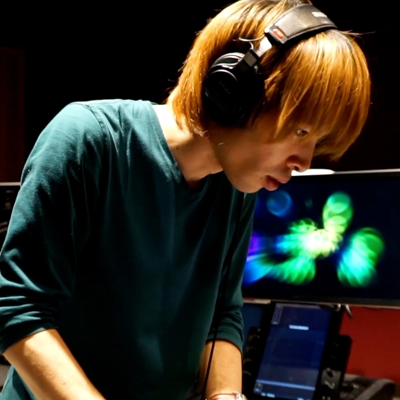 A person with straight, light brown hair wearing headphones and a teal long-sleeve shirt stands in front of monitors, one displaying colorful abstract shapes, focusing intently on equipment, likely in a music studio or DJ booth.