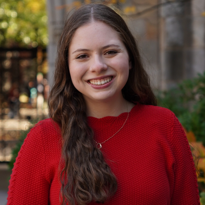 Lilia AitSahlia, a young woman with long, wavy brown hair, smiles at the camera. She wears a bright red sweater and a delicate pendant necklace. Blurred greenery and a stone building exterior set an inviting outdoor scene.