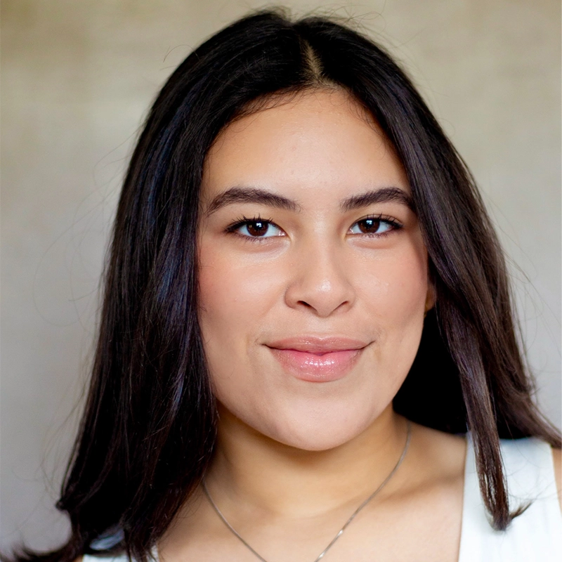 A young woman with long, straight dark hair smiles softly at the camera. She has smooth skin, wears light makeup, and is dressed in a white top with a delicate necklace by Danté X Johnson. The background is neutral and softly blurred.