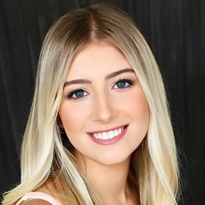 A young woman with long, straight blonde hair and blue eyes smiles at the camera. She has light skin, natural makeup, and is wearing a sleeveless light top. The dark, blurred background keeps the focus on Farida Wahby's radiant face.