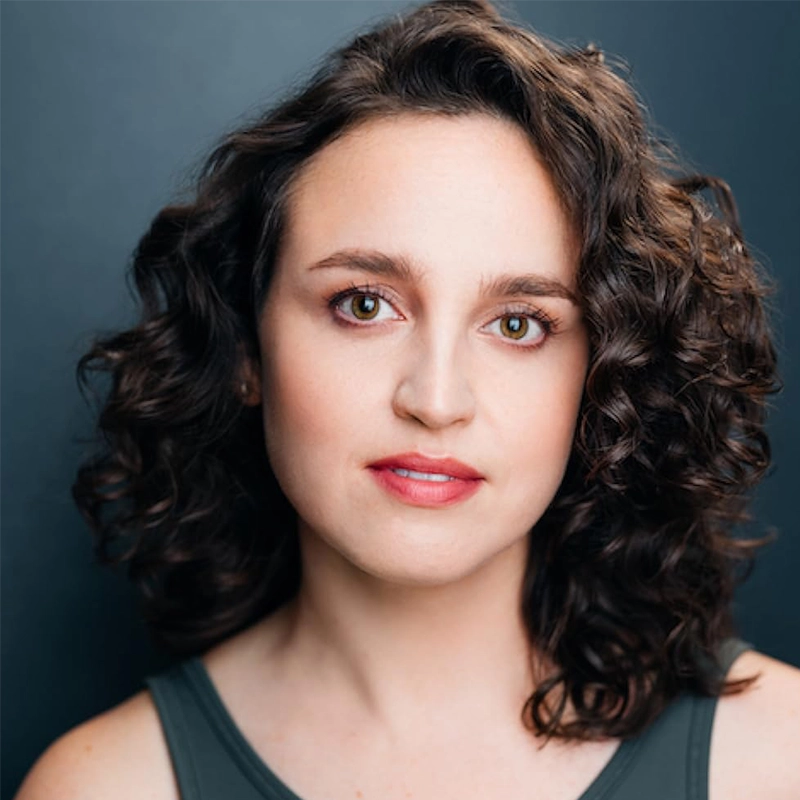 A woman with fair skin and shoulder-length dark brown curly hair faces the camera against a dark background. She has brown eyes, natural makeup, and a slight smile. She wears a sleeveless dark top. The lighting is bright and even on her face.