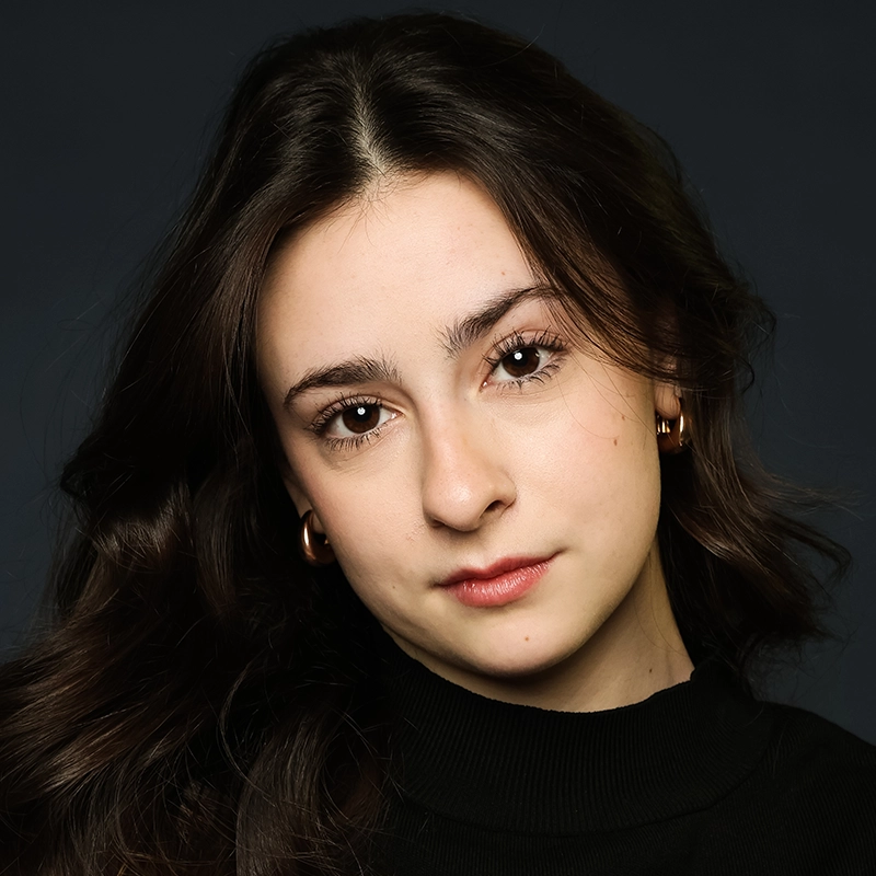 A young woman with long, wavy dark hair and fair skin faces the camera with a neutral expression. Sophia Davison wears a black turtleneck sweater, gold hoop earrings, and subtle makeup, set against a dark, plain background.