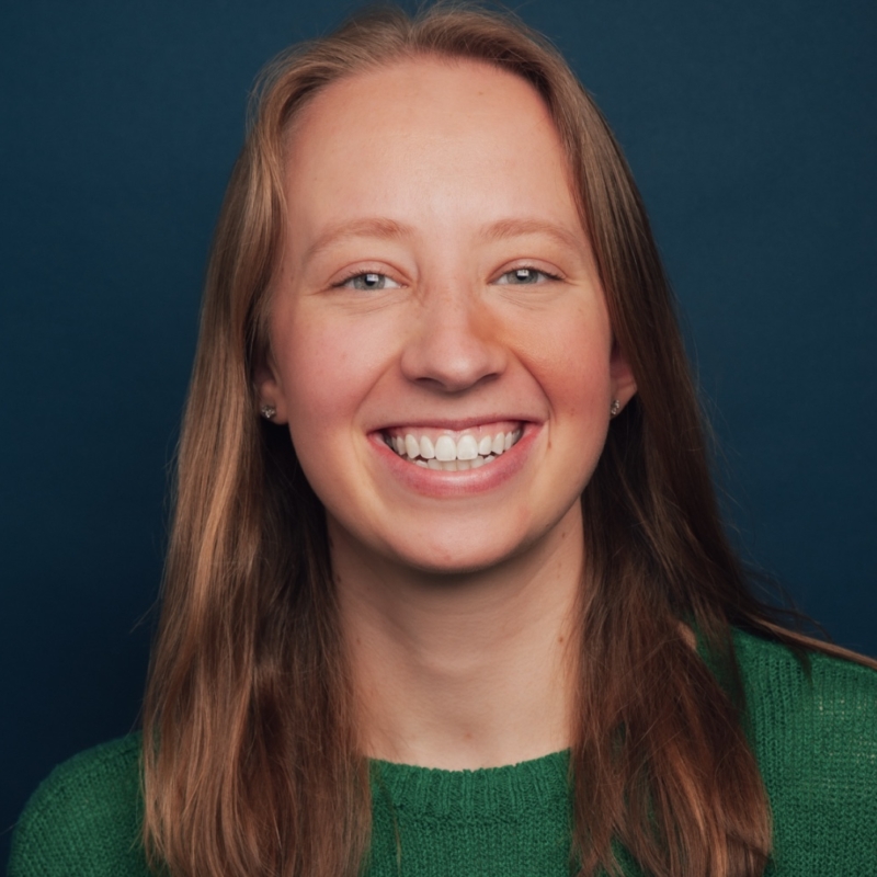 A woman with straight, light brown hair smiles broadly, showing her teeth. She wears a green sweater and small stud earrings. The background is a solid dark blue, and the lighting is bright, highlighting her cheerful expression.