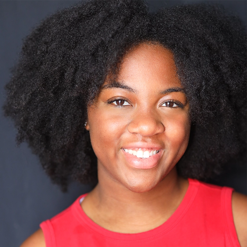 A young Black woman with natural, voluminous curly hair smiles warmly. She wears a sleeveless red top and faces the camera, captured in this Danté X Johnson portrait. The lighting highlights her clear skin and bright expression against a dark background.
