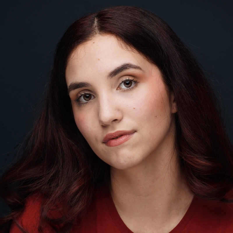 A young woman, Annie Thomas, with long, wavy dark brown hair and fair skin looks directly at the camera. She has brown eyes, natural makeup, and wears a red top. The dark blue background creates a soft, professional portrait feel.