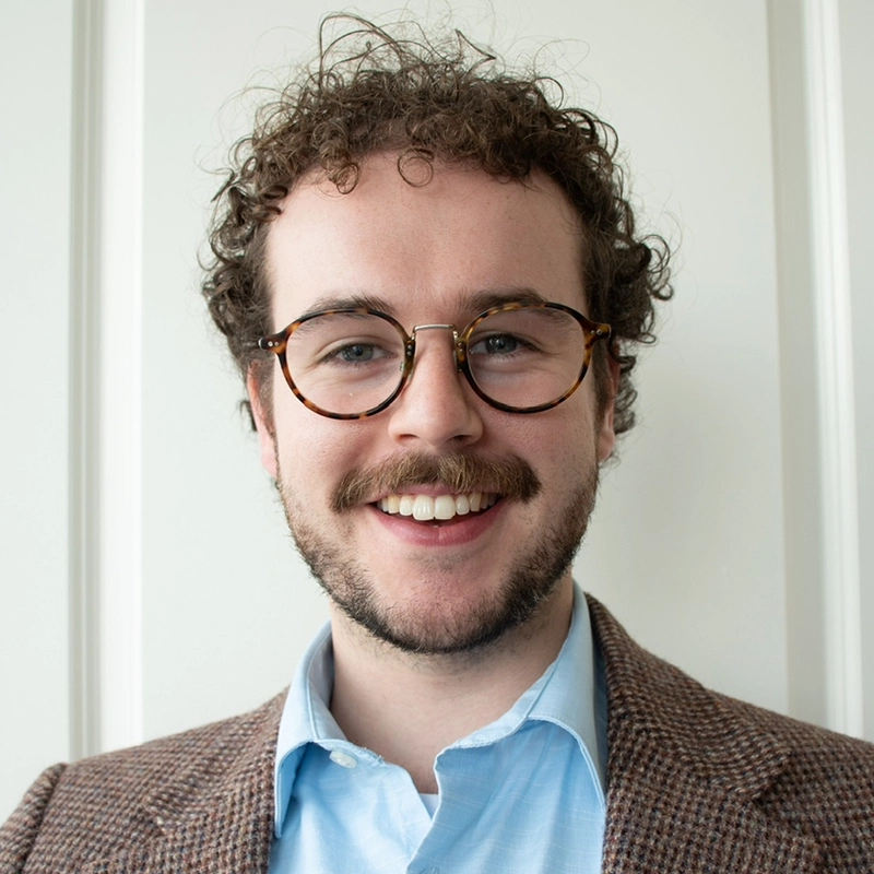 A young man with curly brown hair, a beard, and mustache is smiling. He wears round, brown eyeglasses, a light blue collared shirt, and a brown checked blazer. The background features a plain white wall with vertical panel detailing at Audrey Graves.