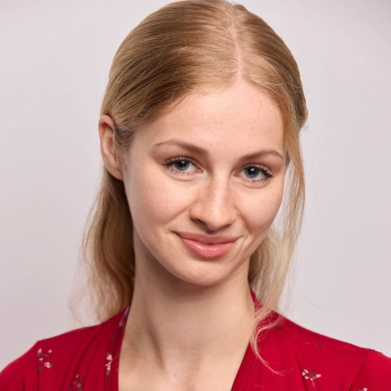 Annie Thomas, a young woman with fair skin and long, light blonde hair parted in the middle and tucked behind her ears, smiles softly. She wears a red top with a subtle floral pattern against a plain, light background that highlights her gentle expression.